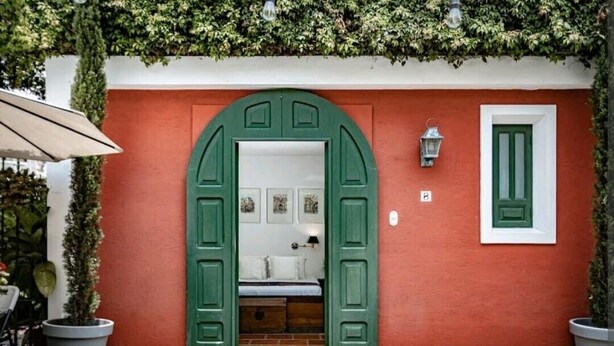 Un edificio rojo con un arco de entrada verde, una cama pequeña en su interior y una planta en maceta a la izquierda.