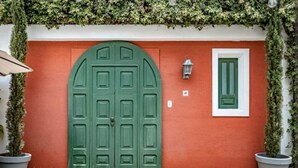 Una puerta verde en arco con marco blanco, una pequeña ventana con persiana verde, y plantas en macetas a cada lado.