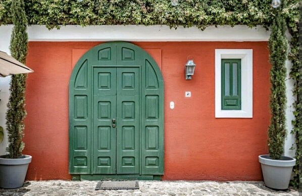Una puerta verde en arco con marco blanco, una pequeña ventana con persiana verde, y plantas en macetas a cada lado.