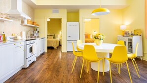 Una cocina moderna con electrodomésticos blancos, un comedor con sillas amarillas y una cama visible al fondo.