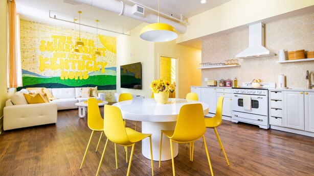 Una sala moderna con un sofá blanco, sillas amarillas y una mesa de comedor. Se observa una cocina con armarios blancos y una estufa.