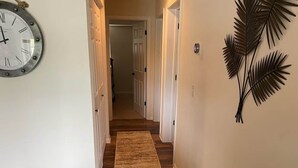 A hallway with a wooden floor, a clock on the left, and a wall-mounted plant on the right.