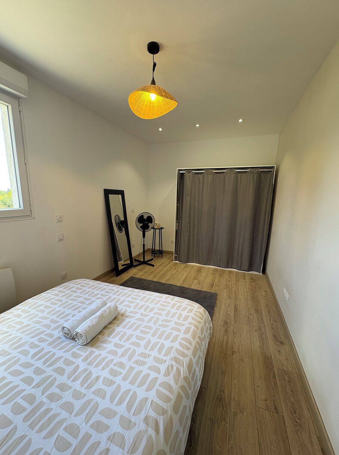 Une chambre moderne avec un grand lit, un ventilateur sur pied, un miroir et une fenêtre.