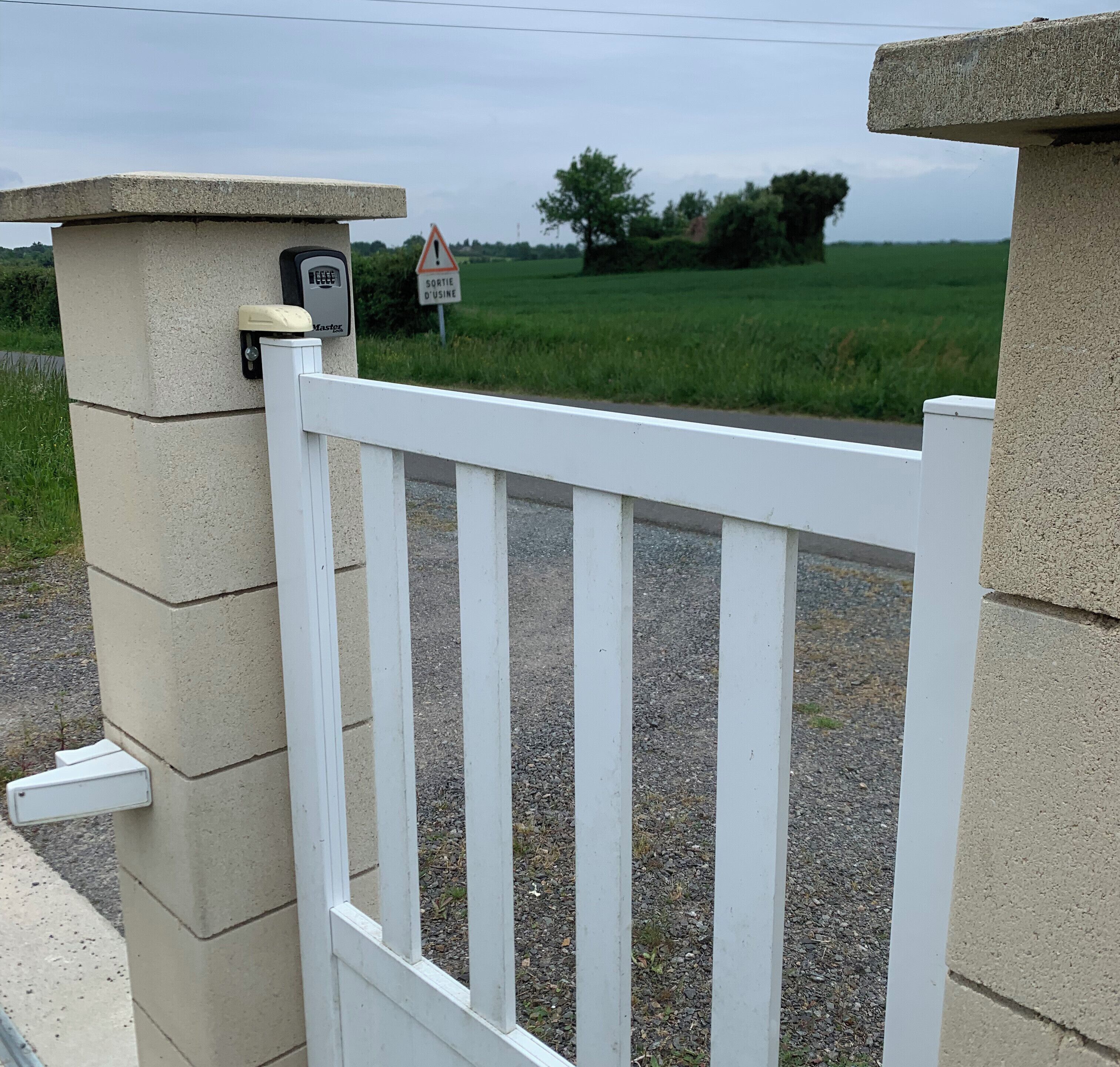 Une entrée fermée par un portail, une clôture en bois peinte en blanc, un panneau d’avertissement et, en arrière-plan, un paysage rural.