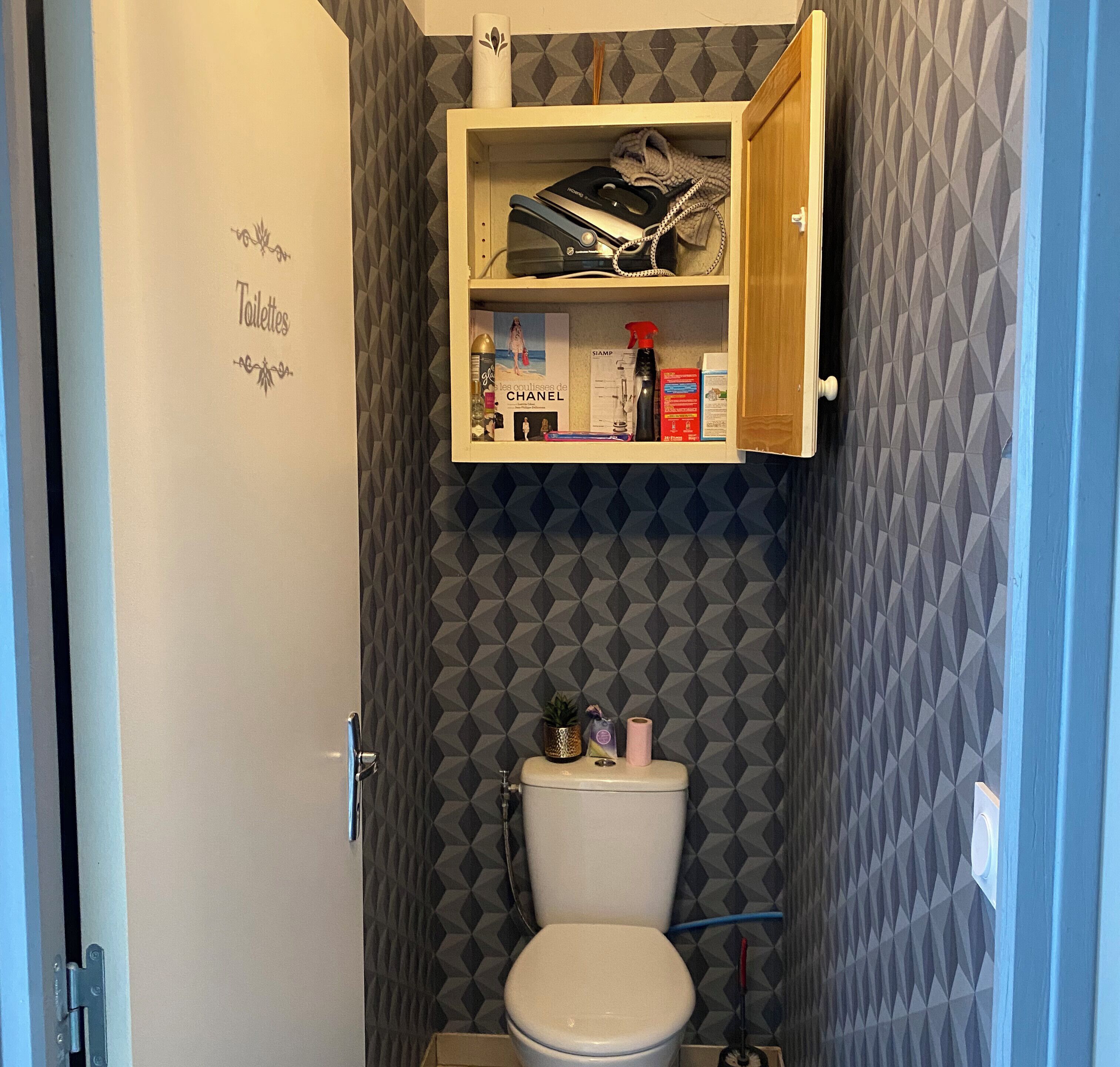 Une petite salle de bain comprenant des toilettes, un meuble au-dessus des toilettes et un mur orné d’un motif géométrique.
