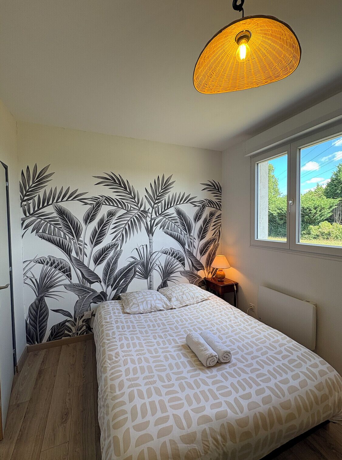 A bedroom with a bed, a nightstand, a lamp, a window, and a wall with a leaf pattern.
