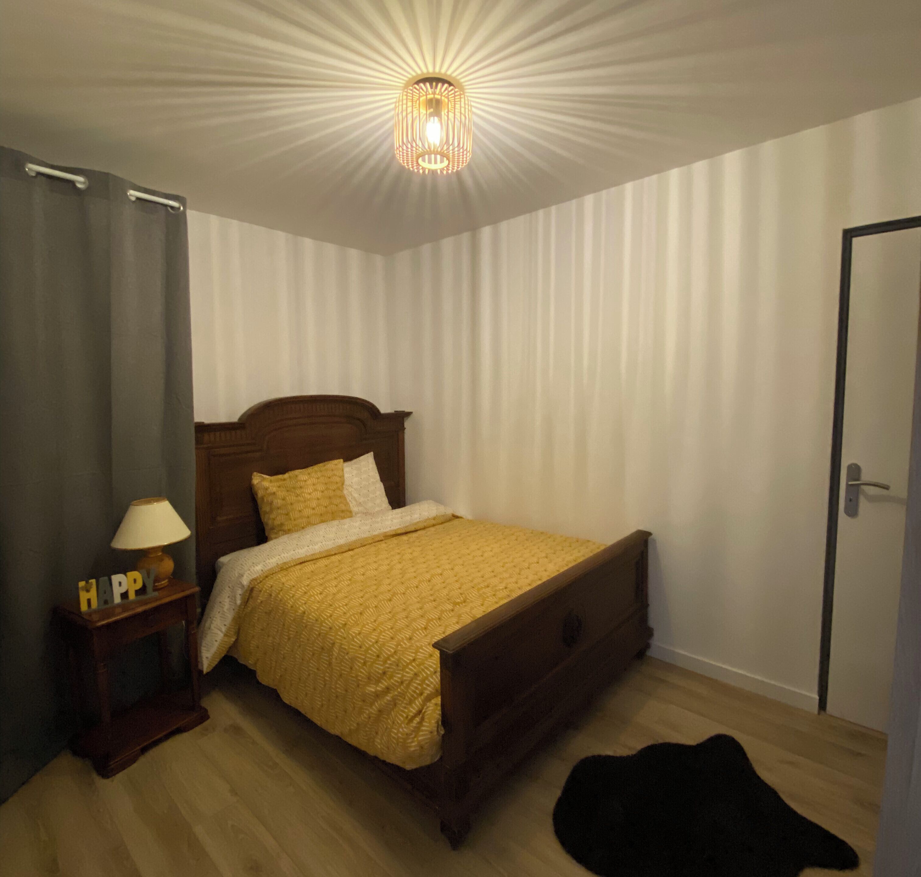 A bedroom with a wooden bed, a bedside table with a lamp, a 'HAPPY' sign, and a black rug on the floor.