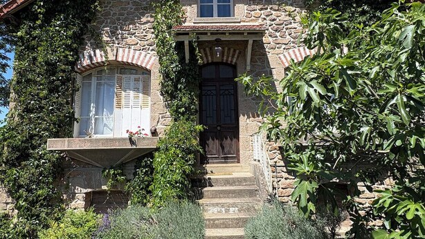 Una casa de piedra con una entrada central, un ventanal sobre la puerta y un pequeño porche.