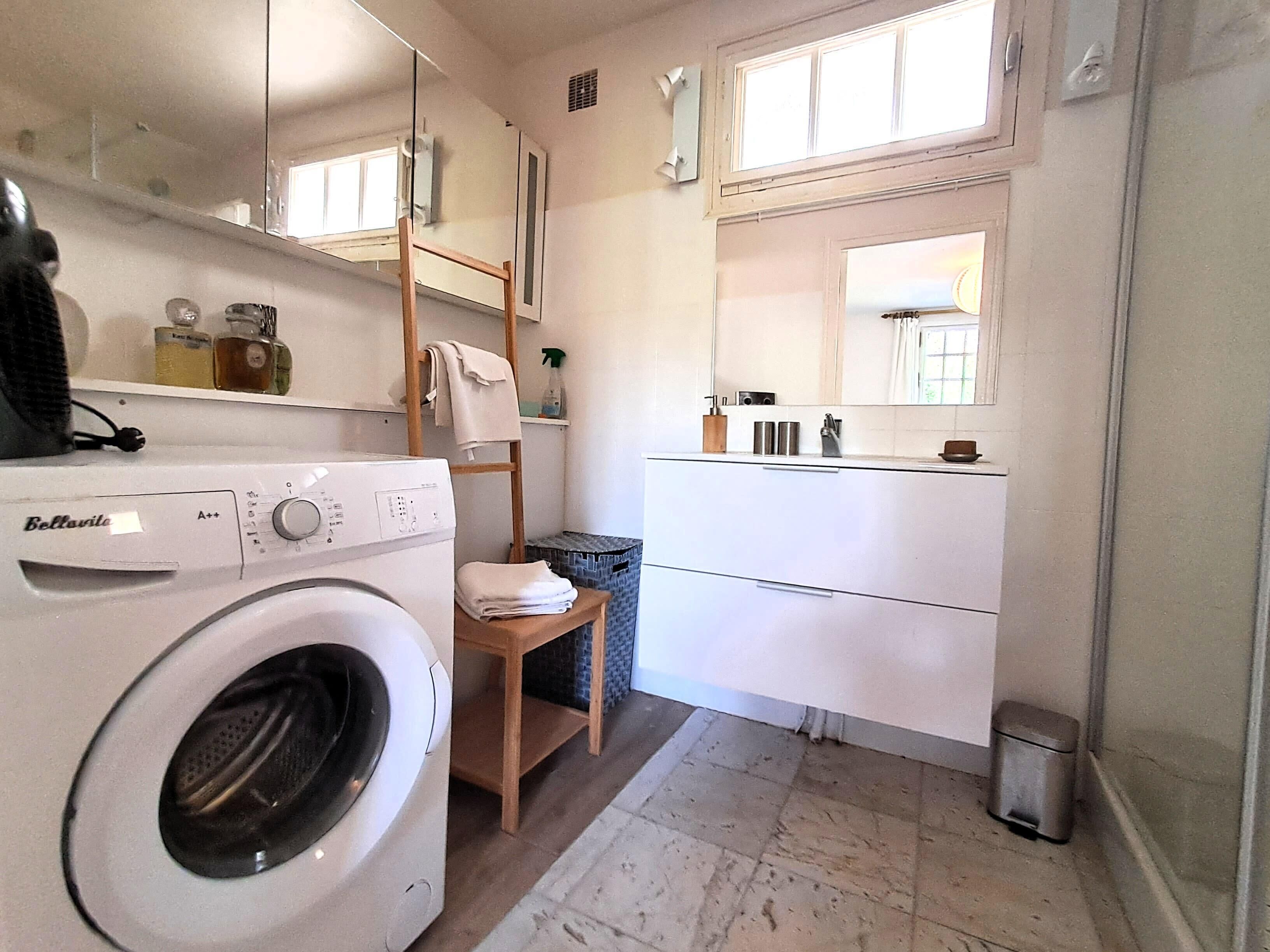 A compact laundry room with a front-loading washing machine, a drying rack, and a small chair.