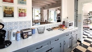 Una cocina con encimera blanca, cafetera, hervidor y tostadora. Hay carteles enmarcados en la pared y una ventana con cortinas.