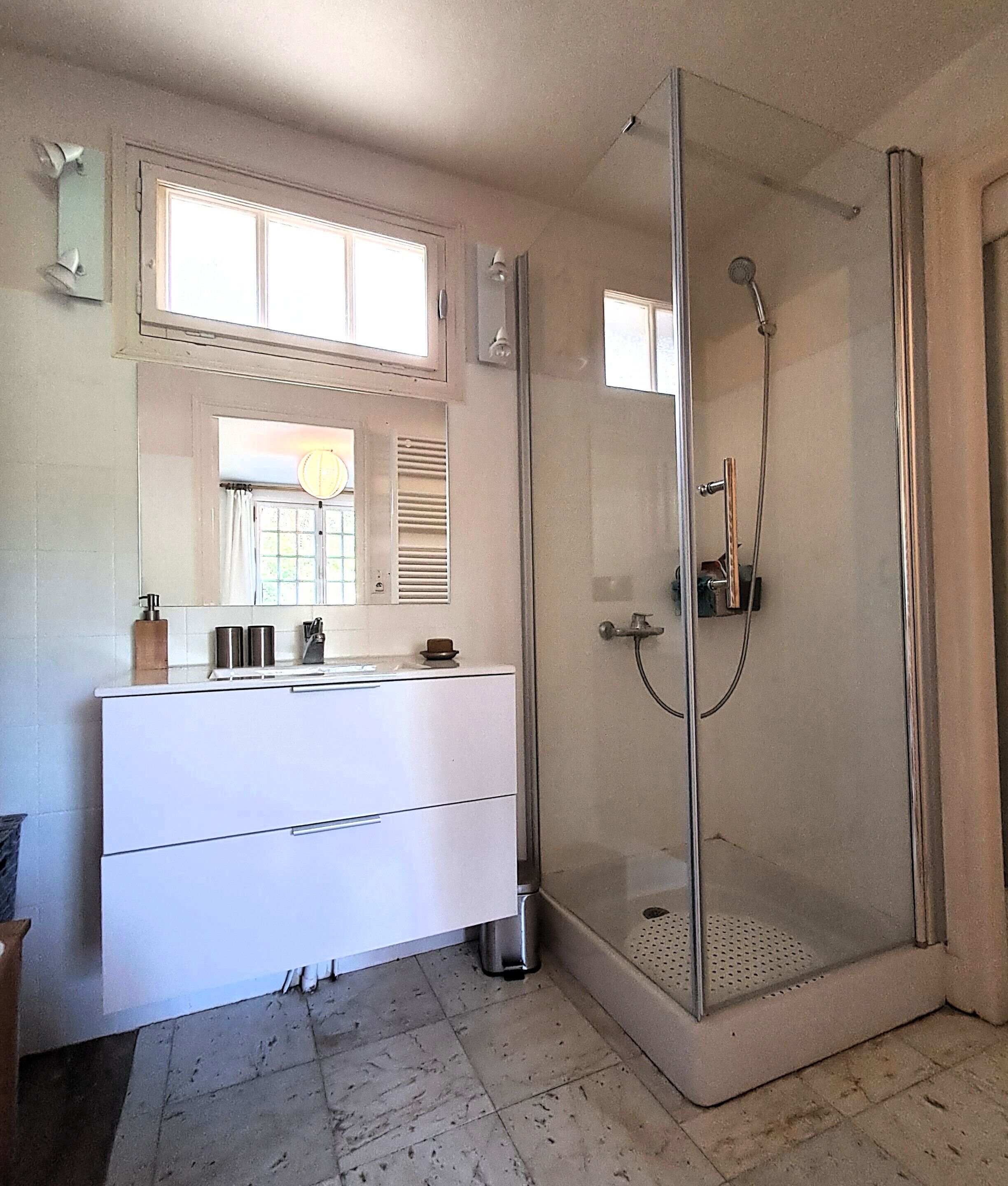 A bathroom with a glass-enclosed shower, a white vanity with a mirror, and a window with blinds.