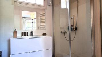 A bathroom with a glass-enclosed shower, a white vanity with a mirror, and a window with blinds.