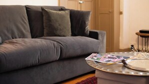 Un canapé gris avec deux coussins, une petite table ronde sur laquelle repose un magazine, et une porte visible au fond.