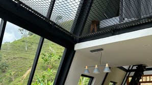 A modern living room with a grey sectional sofa, a dining area with a black table and chairs, and a large window offering a view of lush greenery. - AREWÁ Biohabitat (Pijao)