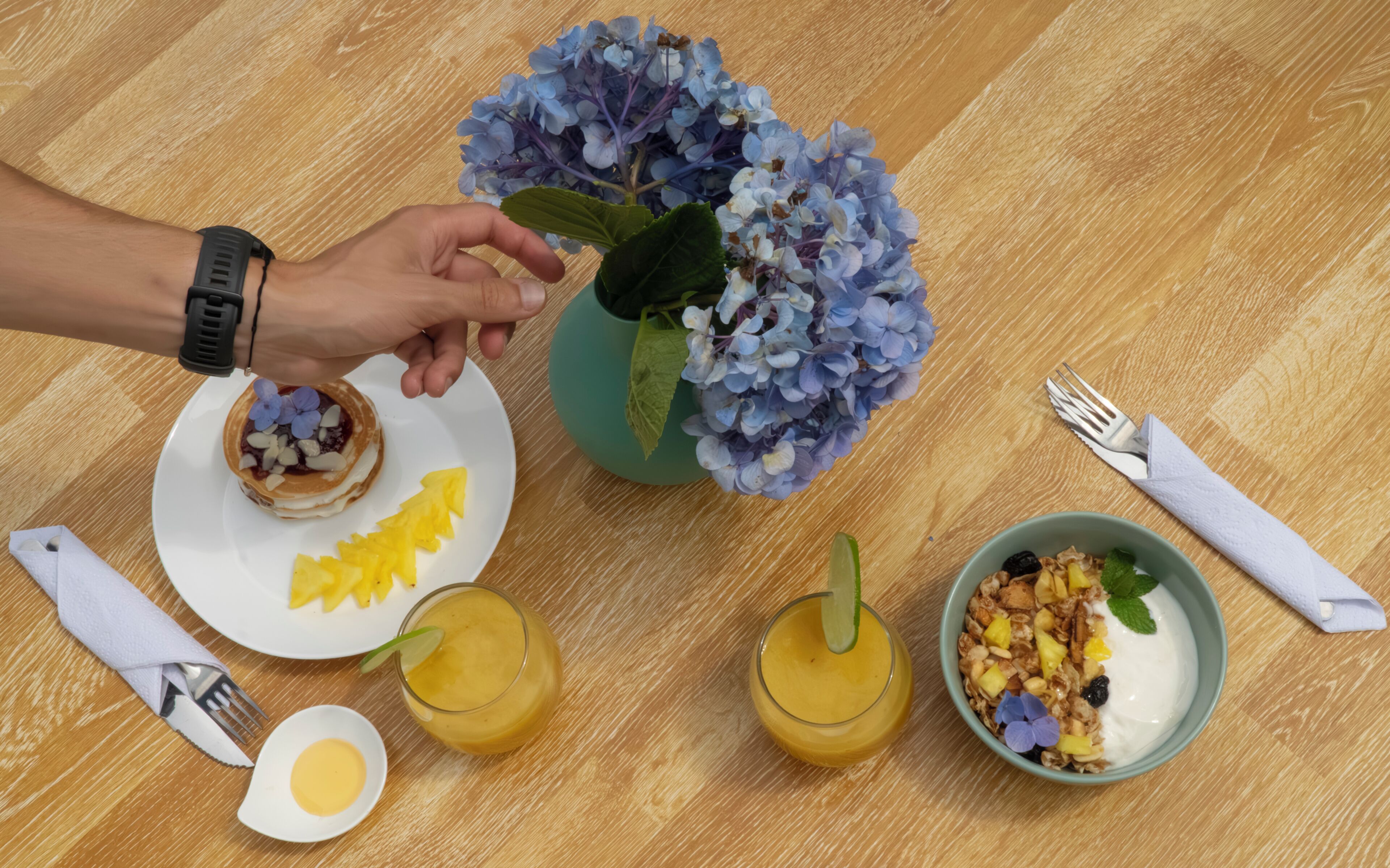 Impostazione colazione con pancakes, insalata di frutta, un bicchiere di succo d'arancia, una ciotola di cereali e un vaso di fiori su un tavolo di legno.