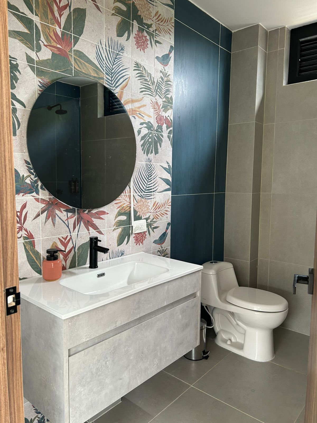 A bathroom with a round mirror, a sink, and a toilet. The walls are decorated with a tropical leaf pattern.