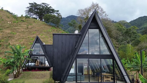 A modern, triangular house with large glass windows, set in a lush, hilly landscape. - AREWÁ Biohabitat (Pijao)