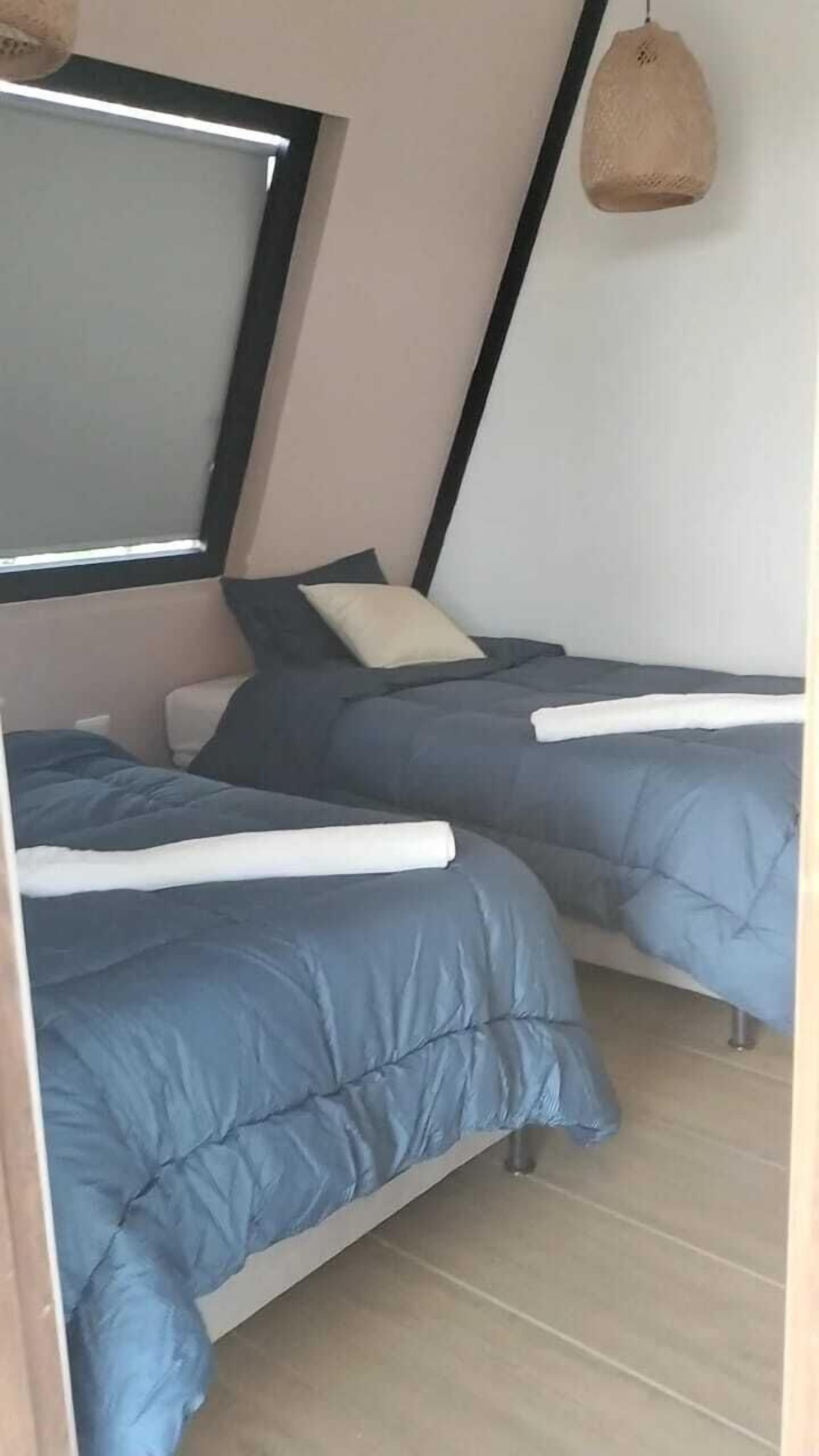 A room with two beds, each with a blue duvet and white pillows, a large window with a black frame, and a woven light fixture.