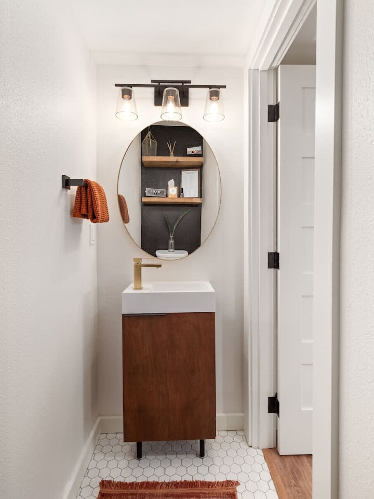 Une salle de bain avec un miroir rond, un lavabo blanc et un meuble en bois. Il y a trois lampes au-dessus du miroir et un porte-serviettes avec une serviette orange.