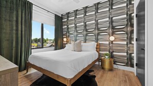 A modern bedroom with a large bed, a wooden headboard, a wall-mounted light fixture, and a view of the outdoors. - 2 Condos-8 Min to Broadway-Balcony-5 King Beds (Nashville)