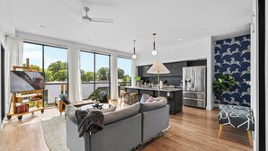 A spacious living area with a grey sofa, wooden flooring, a ceiling fan, and a large window offering a view of trees. - 2 Condos-8 Min to Broadway-Balcony-5 King Beds (Nashville)
