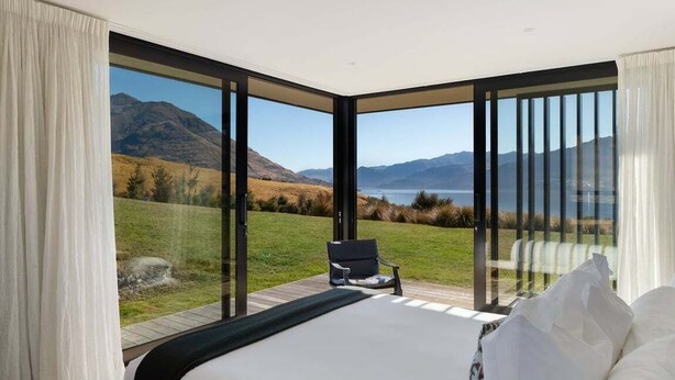 Ein Schlafzimmer mit einem großen Bett, einem Sessel und Blick auf eine gebirgige Landschaft durch raumhohe Fenster.