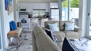 A modern living room with a beige sofa, a bench, and a coffee table. There is a kitchen area with white cabinets, a microwave, and a refrigerator. The room has a large window providing natural light and a view of greenery outside. - Two private beachfront houses, sleeps 12, kayaks, private heated pools (Cabo Rojo)