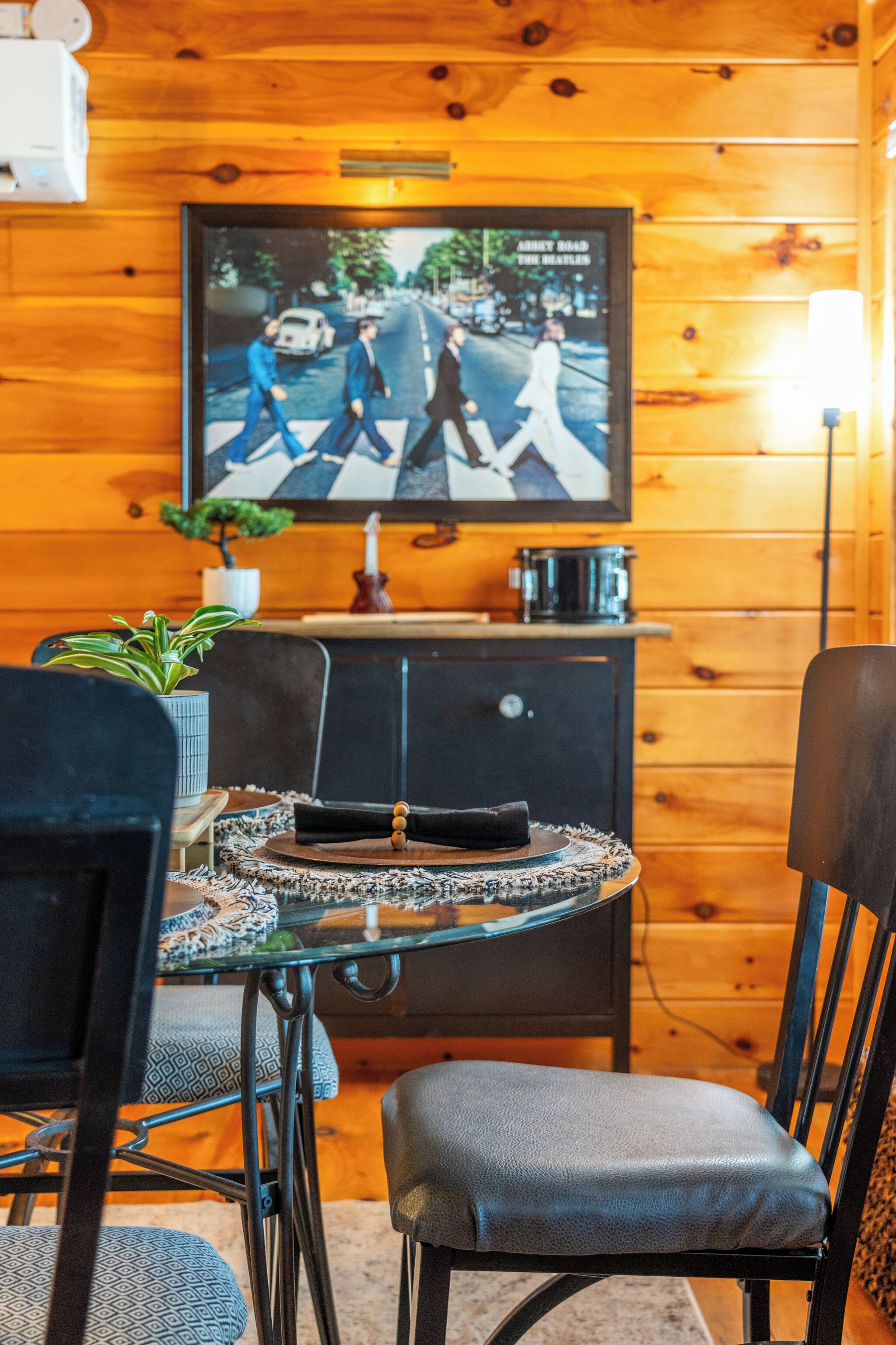 Un interior de madera con un televisor mostrando 'Abbey Road' de The Beatles, una mesa de comedor con sillas y una pequeña planta en maceta.