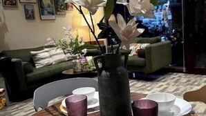 A dining area with a table set for two, a vase with white flowers, and a green sofa in the background.