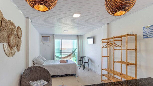 A hotel room with a bed, a wicker chair, a wooden shelf, and a view of the outdoors. - Loft com Varanda e Vista Para o mar em Mangaratiba (Mangaratiba)