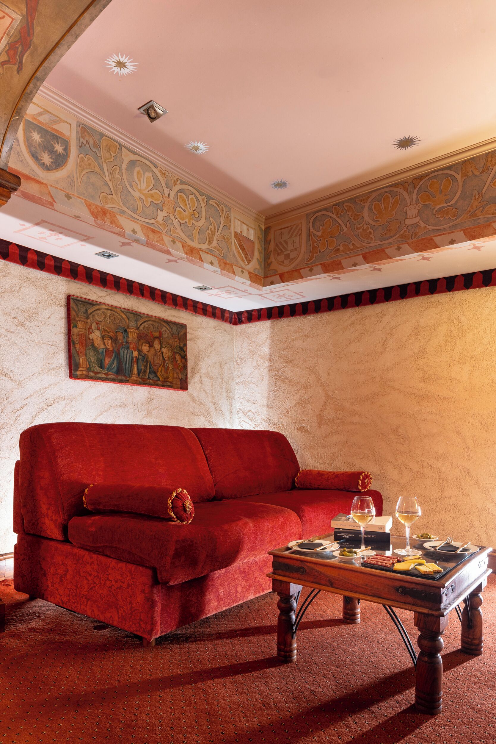 Una habitación con un sofá de terciopelo rojo, una mesa de centro de madera con comida y bebidas, y un mural en la pared.