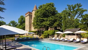 A swimming pool with lounge chairs and umbrellas, a stone tower, and lush greenery. - Château de Codignat (Bort-l'Étang)