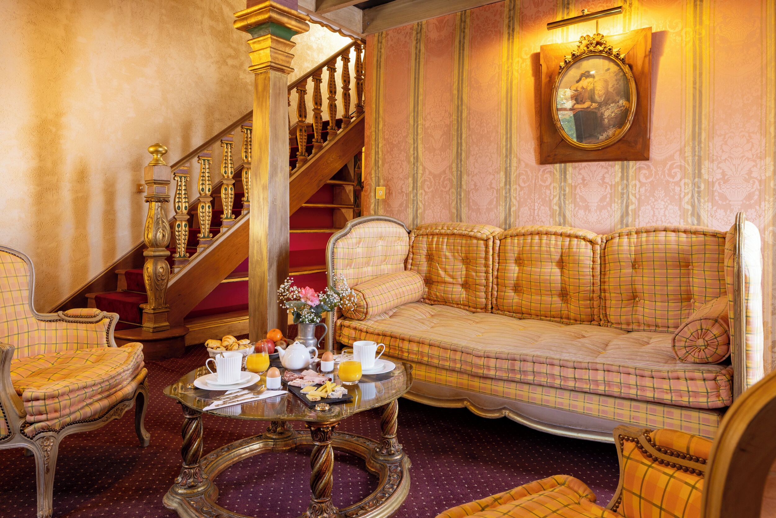 A room with a checkered sofa, a dining table with breakfast items, a staircase, and a wall-mounted mirror.