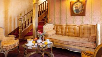 A room with a checkered sofa, a dining table with breakfast items, a staircase, and a wall-mounted mirror.