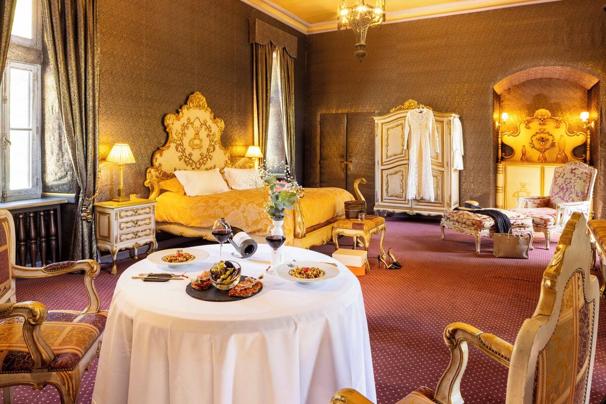 Un dormitorio lujoso con una cama grande, muebles ornamentados y un rincón de comedor con una mesa dispuesta para dos.