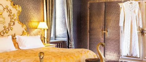 A luxurious hotel room with a large bed, ornate headboard, a vanity with a mirror, and a wardrobe.