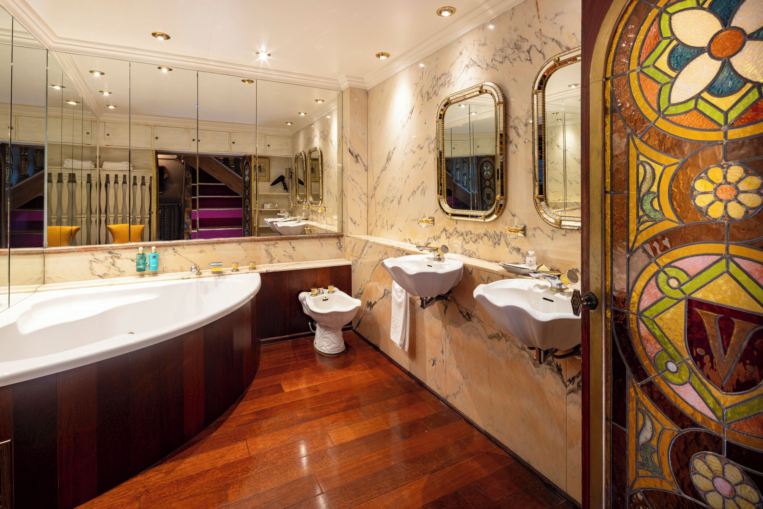 A luxurious bathroom with a large freestanding bathtub, two sinks, a bidet, and a mirror with a stained glass design.
