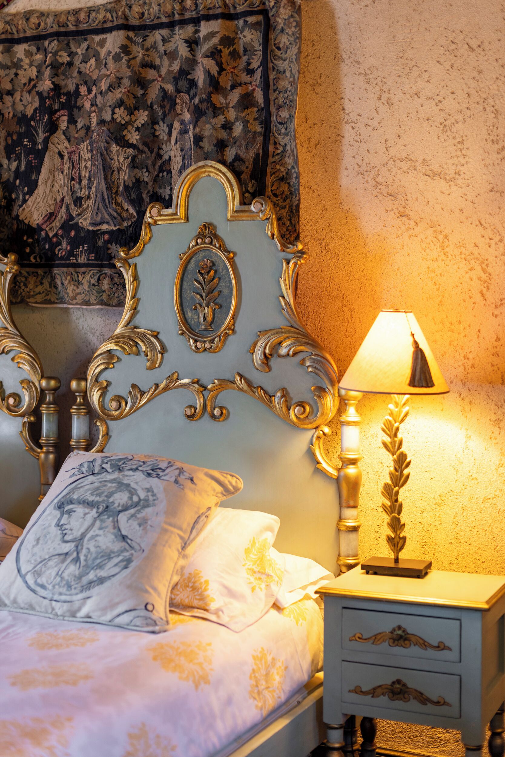 Un lit avec une tête de lit décorée, une table de chevet avec une lampe et une tapisserie au mur.