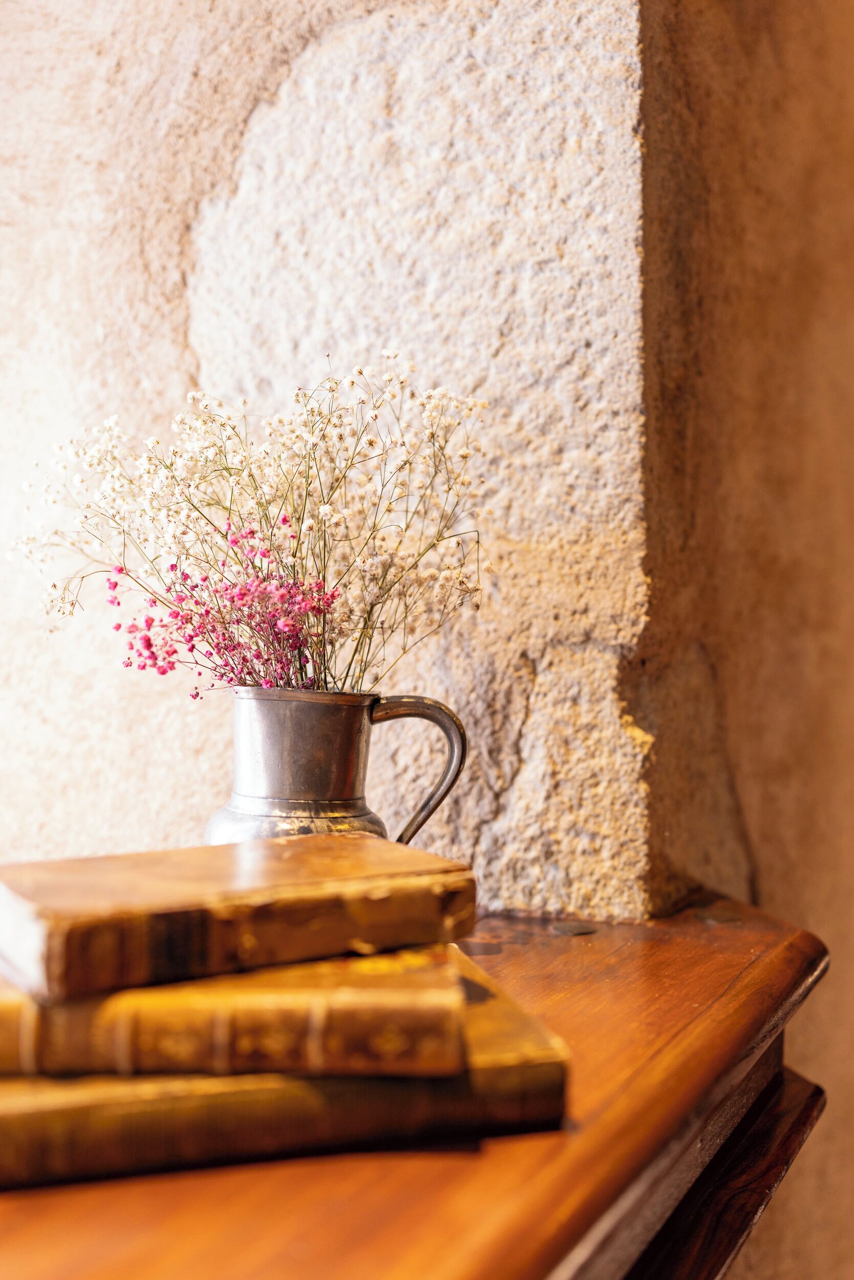A metal pitcher with flowers on a wooden surface against a textured wall.