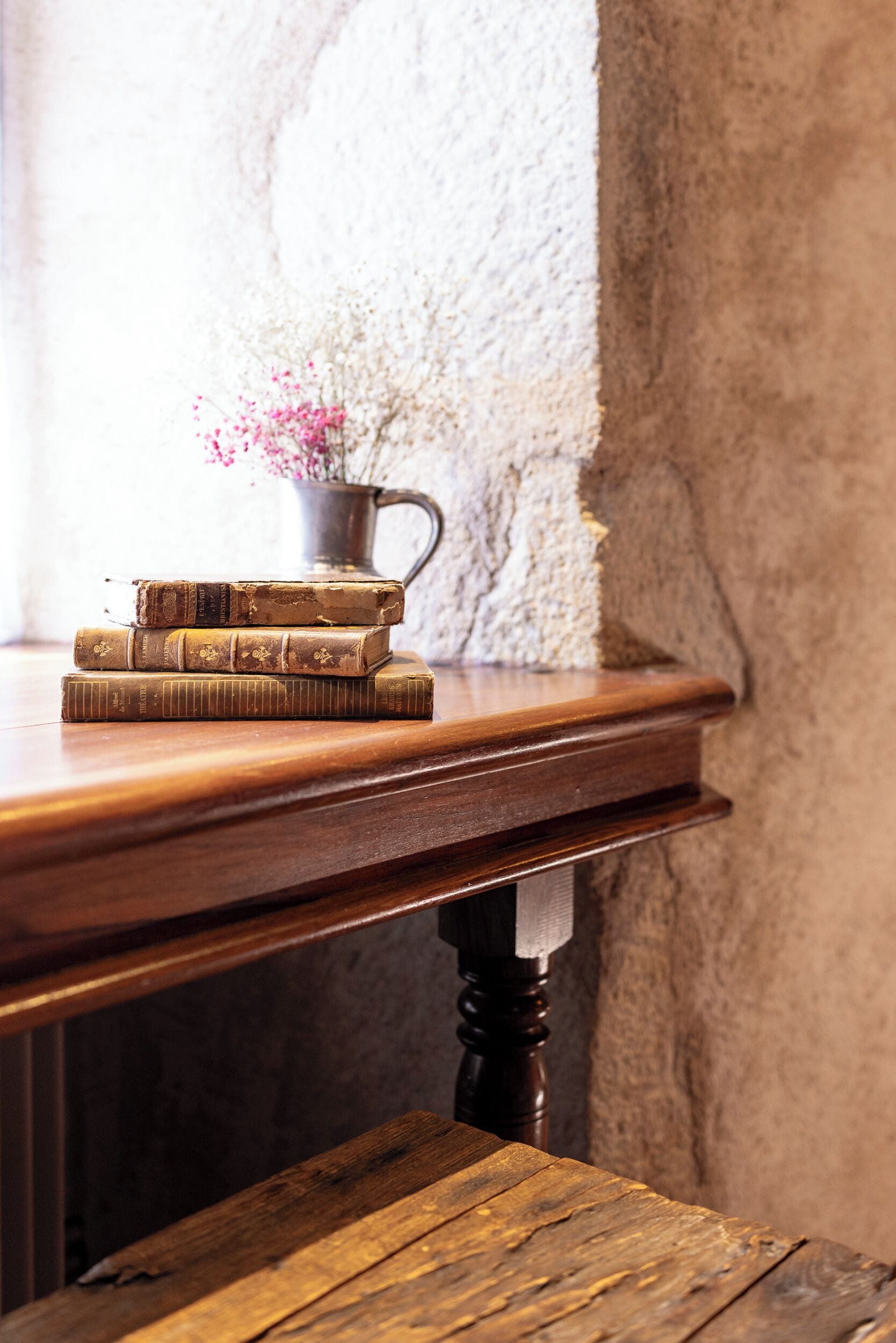 Een houten tafel met een stap oude boeken en een metalen kruik met bloemen.