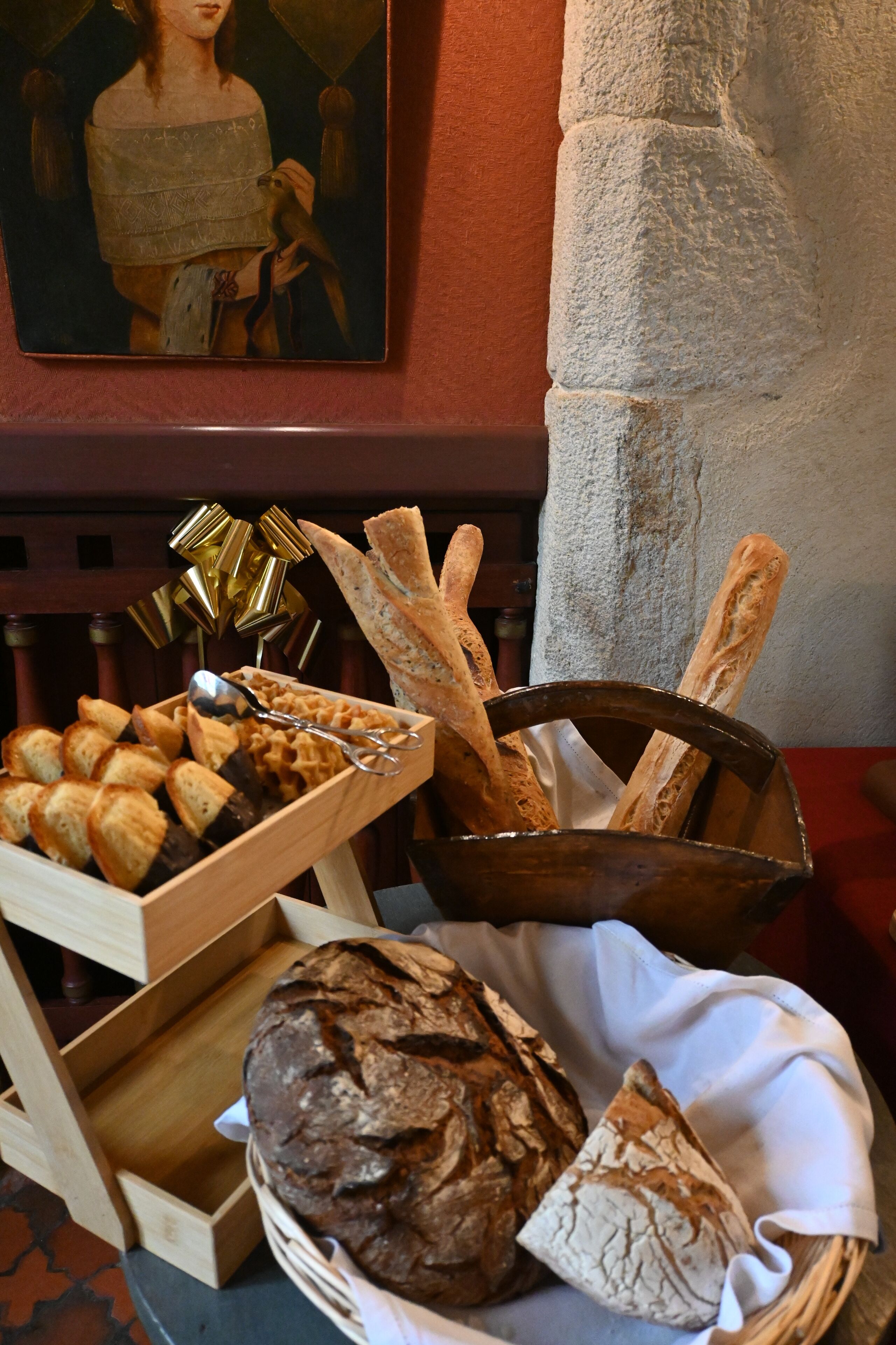 A wooden bread basket with various types of bread and pastries, a wooden tray with more bread, and a framed painting on the wall.