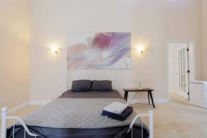 Un dormitorio con una cama, un cuadro abstracto colgado en la pared, lámparas de noche, una mesita de noche y una puerta.