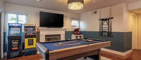 A room with a pool table, a flat-screen TV, arcade games, and a fireplace.