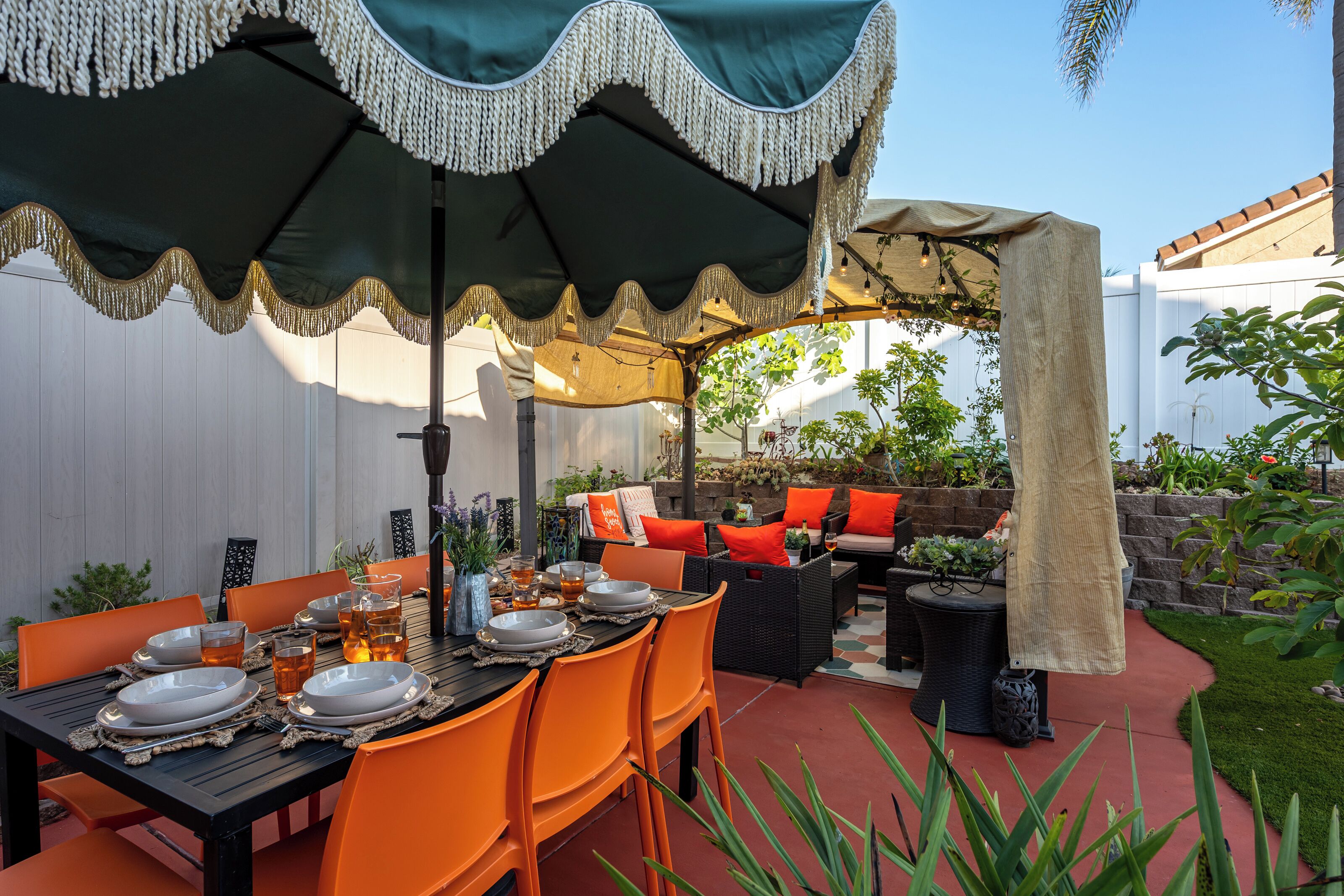 An outdoor patio with a dining table set for four, surrounded by orange chairs, under a large umbrella with fringe detailing.