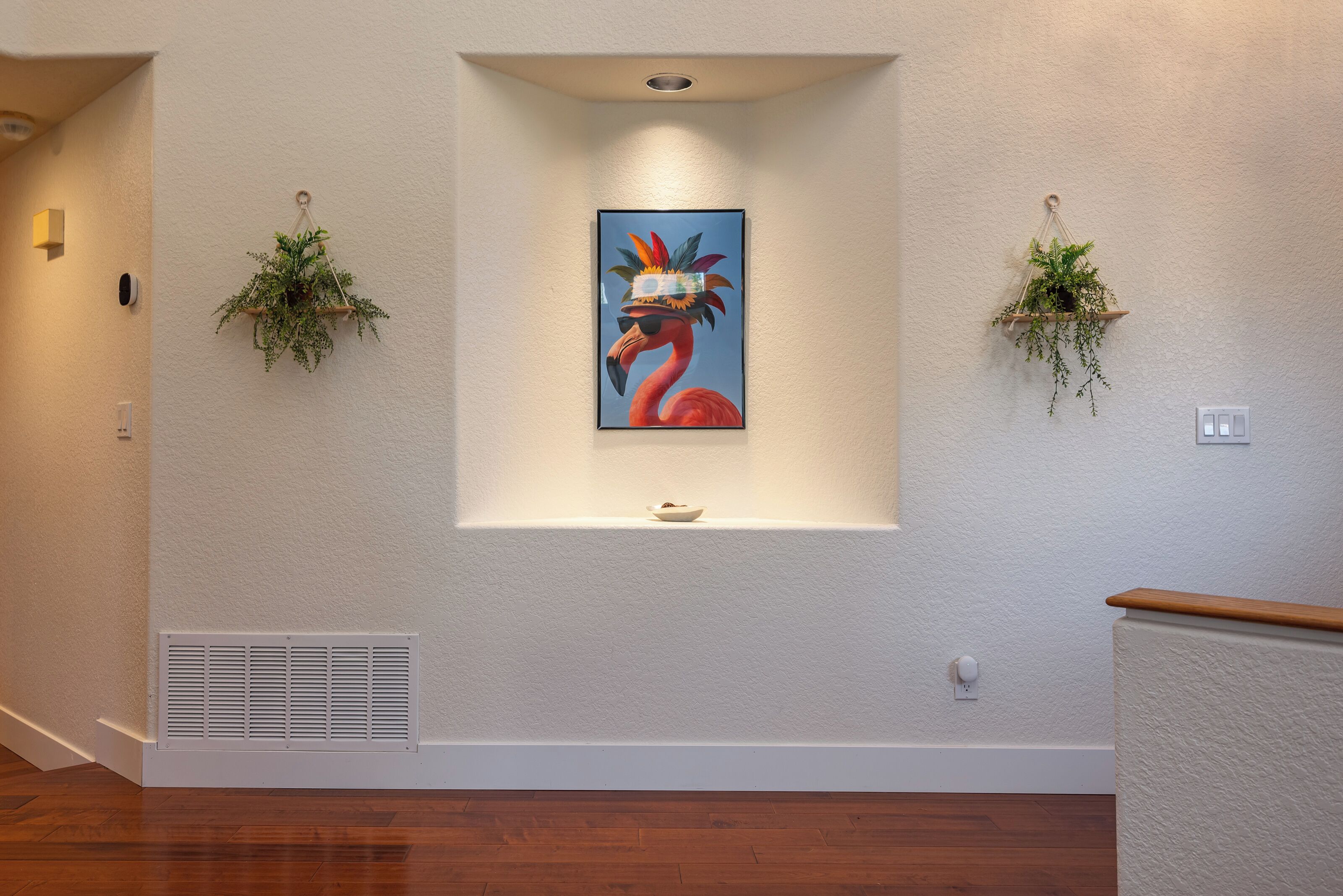 A hallway with a framed painting of a flamingo, two hanging plants, a vent, and a light switch on the wall.