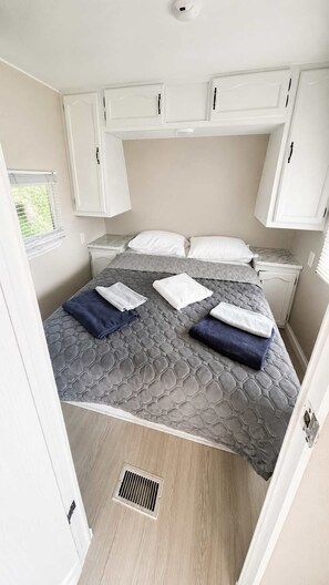 A compact bedroom with a bed, two pillows, a window with blinds, and white cabinets. - Mill Lake Waterfront RV Retreat | Parry Sound #01 (Parry Sound)