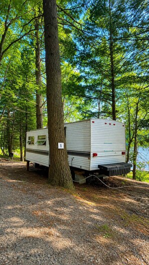 Un camping-car blanc est garĂ© dans une zone boisĂ©e, dâoĂč lâon a une vue dĂ©gagĂ©e sur les arbres environnants et sur une Ă©tendue dâeau en arriĂšre-plan.