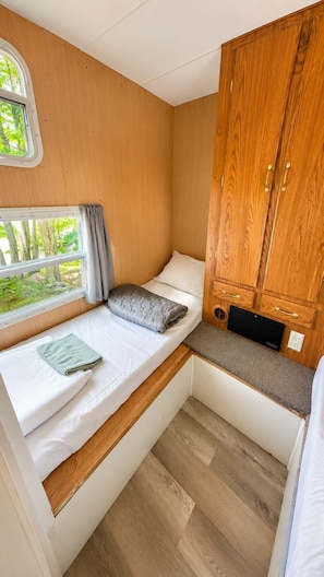 A compact RV interior with a bed, a small seating area, a wooden cabinet, and a window with a view of greenery.