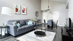 A modern living room with a grey sofa, a coffee table, a TV stand, and a dining area with chairs and a table.