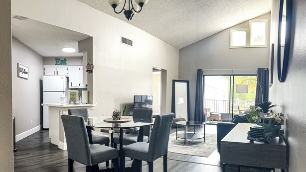 A modern kitchen with white appliances, a dining area with a round table and chairs, and a view of the outdoors through a sliding door.